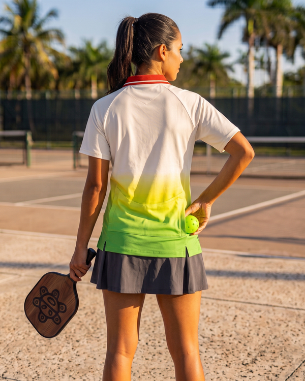 A woman on a pickleball court wearing a white, yellow, and green gradient t-shirt, a gray pleated skirt, and holding a paddle and retrieving a pickleball  ball from her back pocket.